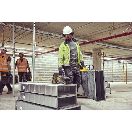 Construction worker using a Grabo 18V XR Suction Lifter to carry metal duct framework, case in other hand and workers in background