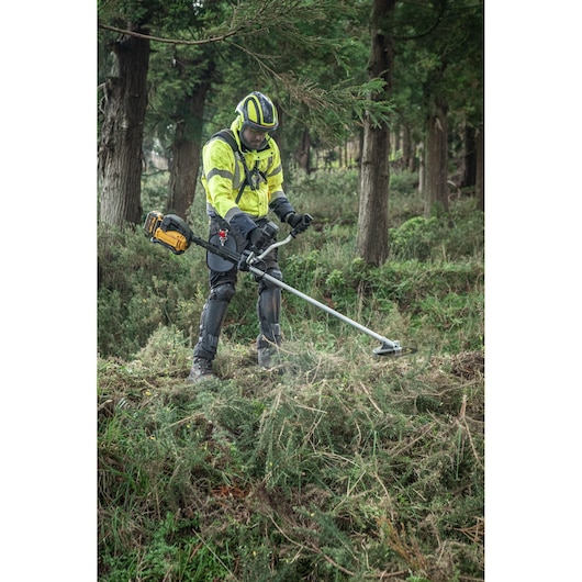 Landscaper in forest wearing full safety gear including a high visibility jacket, helmet with face visor, knee, shin and leg covers. and gloves using the DEWALT 54V Brush Saw (Bull Handle) with harness to cut through dense grass area.
