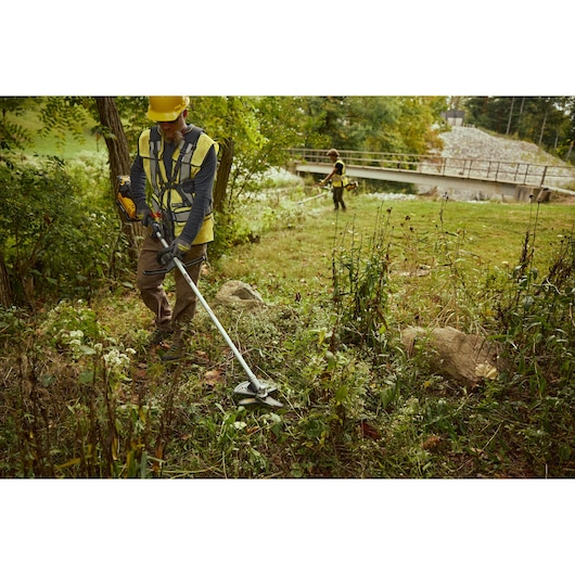 Landscaper using the DEWALT 54V Brush Cutter  (D-Handle) with shoulder strap harness, helmet and gloves in grassy area by river bank.