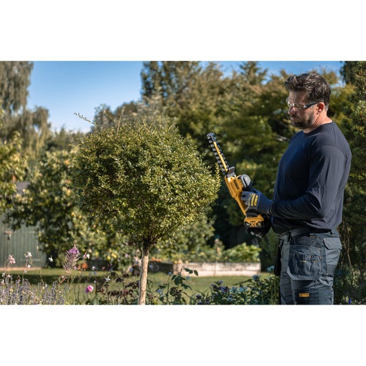 Worker using the DEWALT 18V XR Compact Hedge Trimmer to trim a small tree
